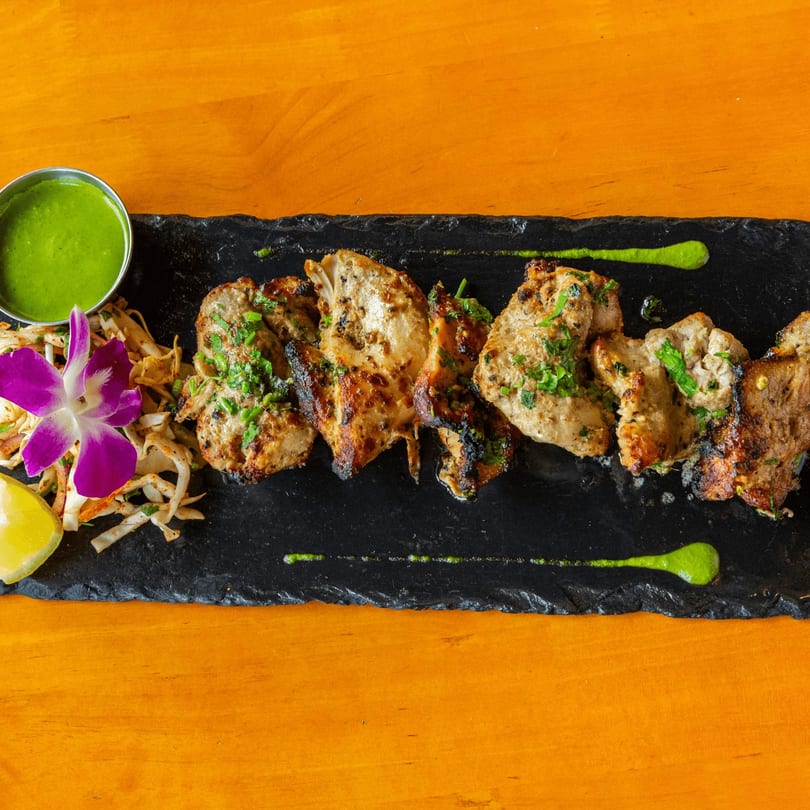 Grilled chicken pieces with green sauce, shredded salad, lemon, and an orchid on a black slate.