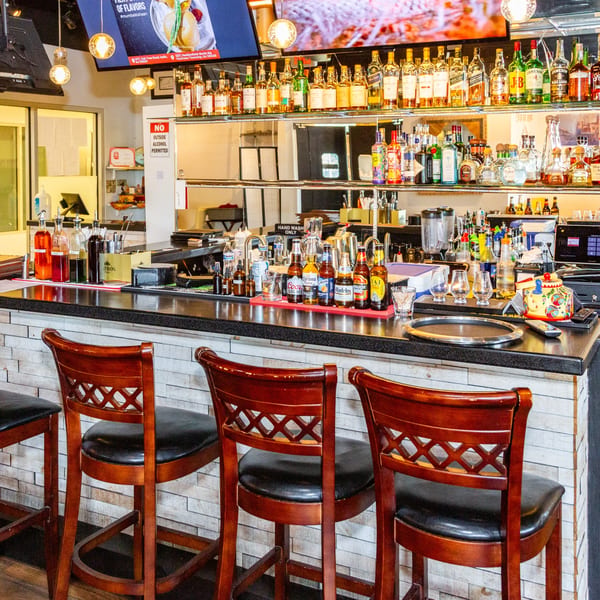 A well-stocked bar with various liquor bottles, beer, and bar stools, under a large TV screen.