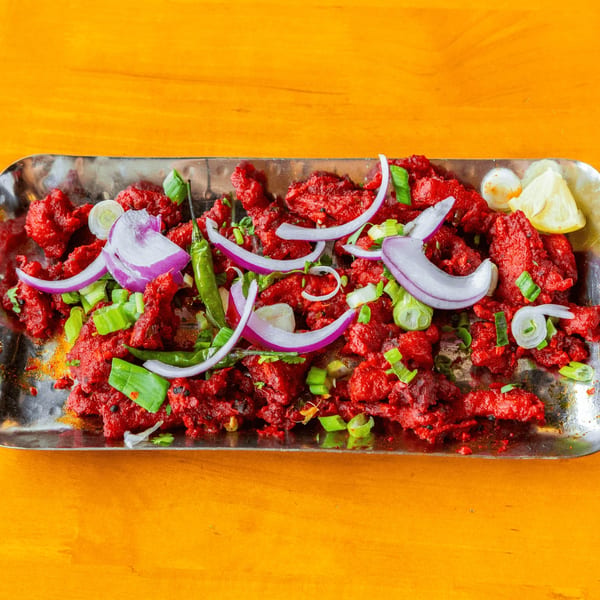 Red chicken dish garnished with sliced red onions, green chilies, and scallions on a metal tray.
