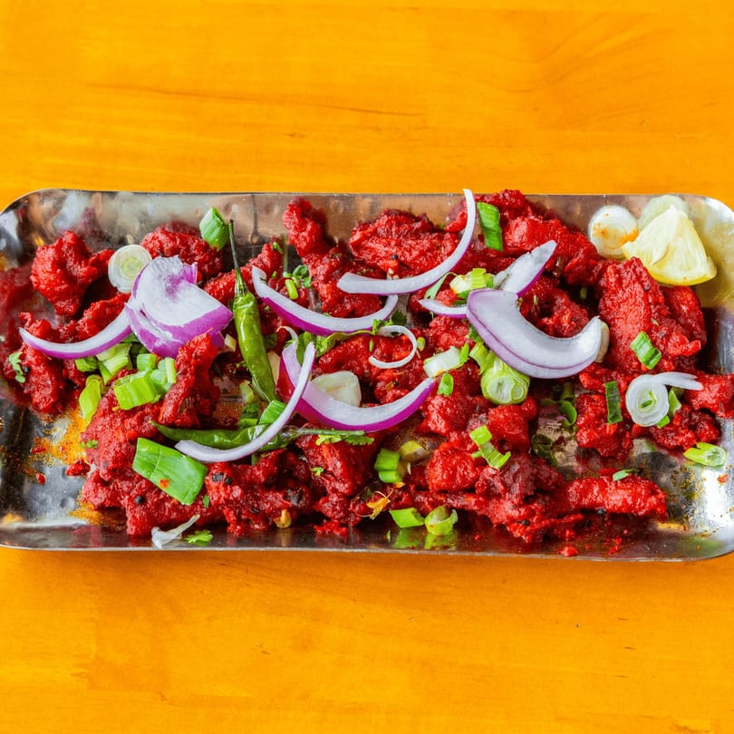 Red chicken dish garnished with sliced red onions, green chilies, and scallions on a metal tray.