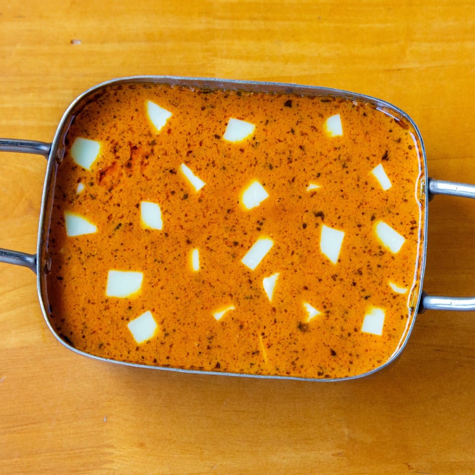 Overhead view of a rectangular metal dish filled with orange curry and white paneer cubes.