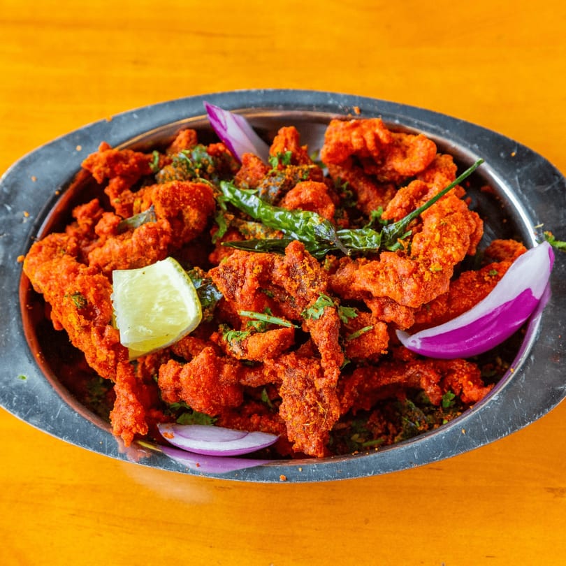 A metal platter filled with bright red fried chicken, garnished with lime, green chilies, and red onion.