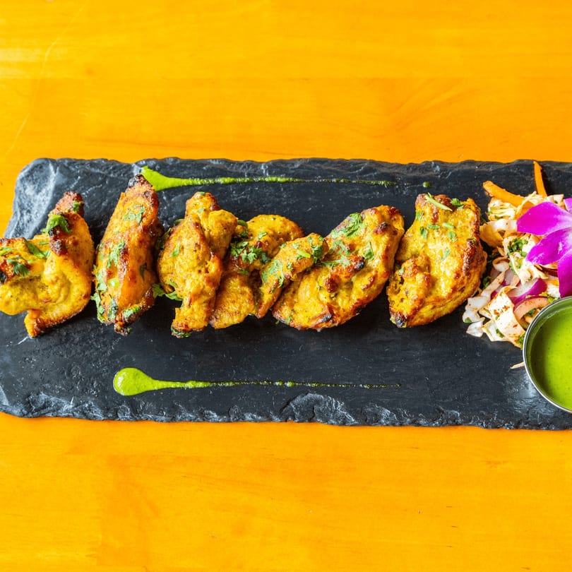 Grilled chicken pieces with green sauce, a side salad, and a purple orchid on a black slate.
