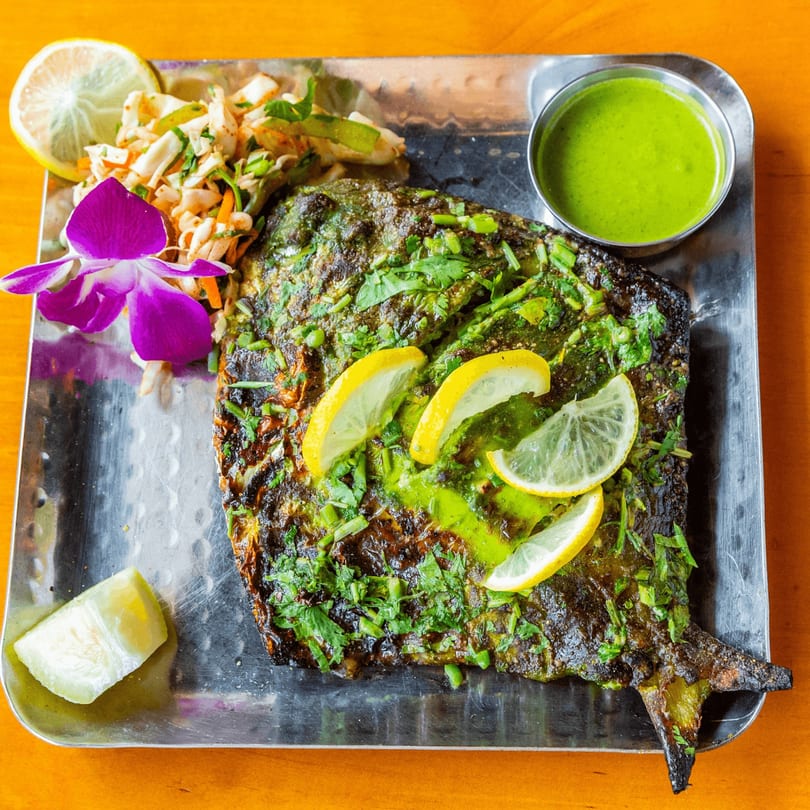 Grilled fish with green sauce, lemon slices, salad, and a purple orchid on a metal tray.